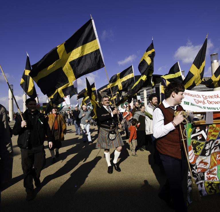 A band marches with flags of St David (which are black with a yellow cross).