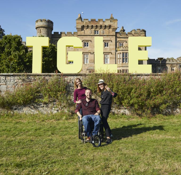 Large letters TGLE in front of a Hawarden castle. 2 females stood and one male sat in a wheelchair in the foreground