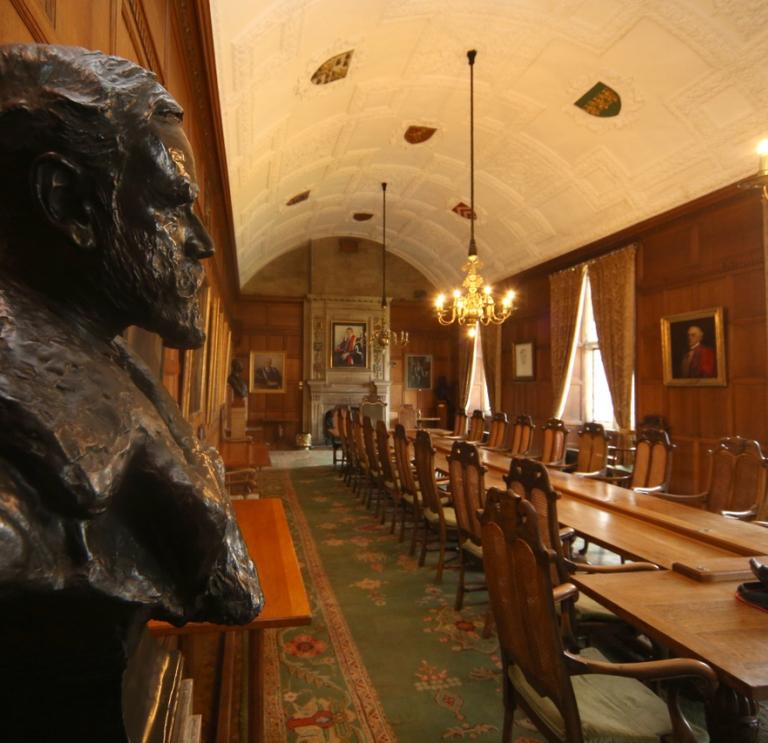 The council chamber witha   head statue of a man low chandelier lights over a log table with fireplace at the end.