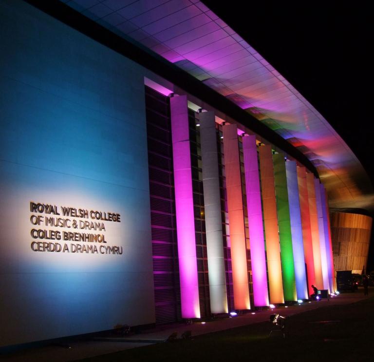 Nightime photo of the exterior with venue name in white lighting and multi colour vertical lit striped down the main building frame. 