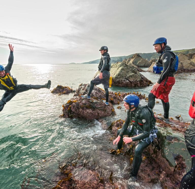 group of male adults laughing as they jump off the rocks into the blue sea, coasteering with instructors.