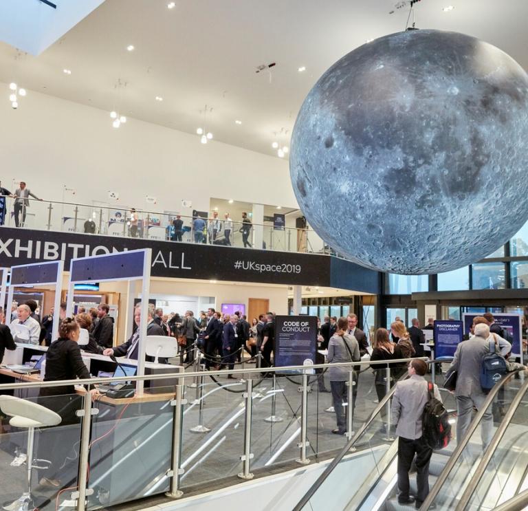 Exhibition hall with people coming up esculators to reception desks and a large grey space globe suspended from the ceiling.