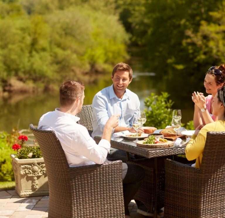 two men and two women in smart cauusal clothing sitting at a table with food and drink outside by the water with red flowers and green trees in the background. 