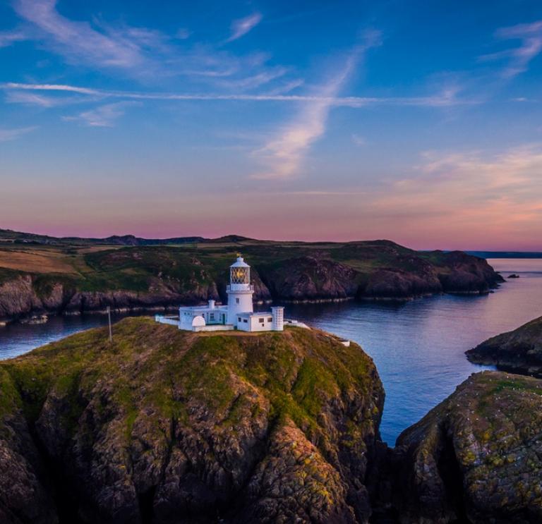 A view of a white lighthouse on a cliff surrounded by the sea with pink and blue sky
