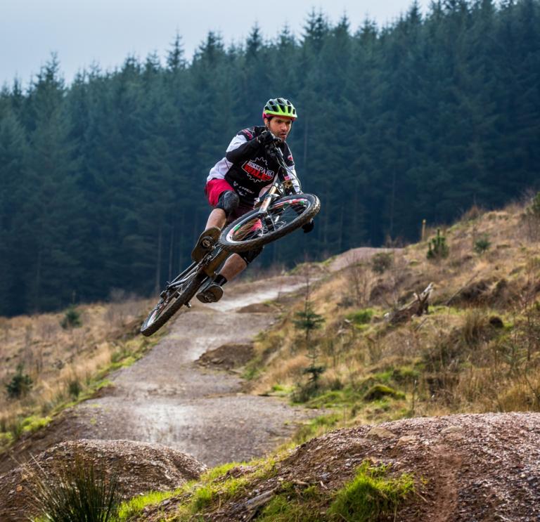 male rider on a mountain bike, airbourne jumping from on a gravel trail, with a forest of trees