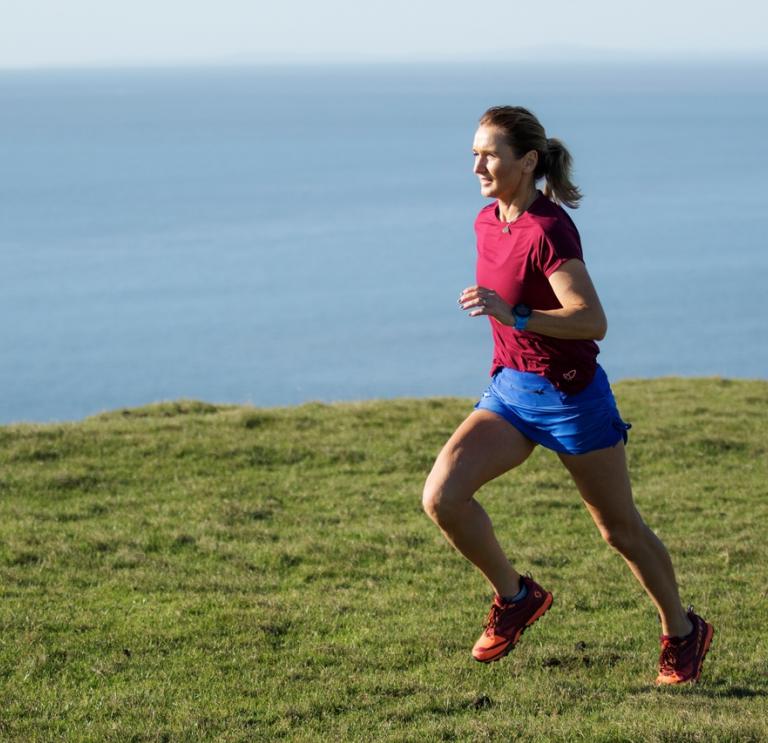 A woman wearing a red top and blue shorts running on grass with the sea in the background