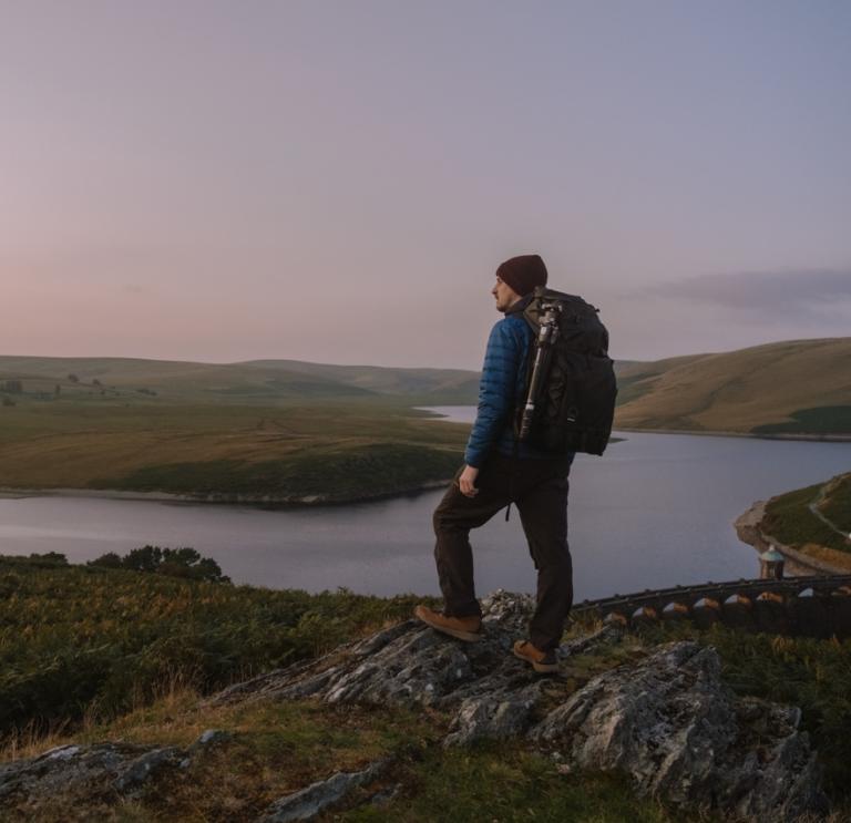 Ein Mann mit einem Rucksack auf dem Rücken steht am Rande eines Berges und blickt auf eine Landschaft aus Bergen und einem See.