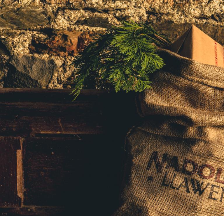 Brown hessian sack stuffed with christmas gifts and printed with the Welsh words Nadolig Llawen, hanging on a wooden mantle against a stone wall.