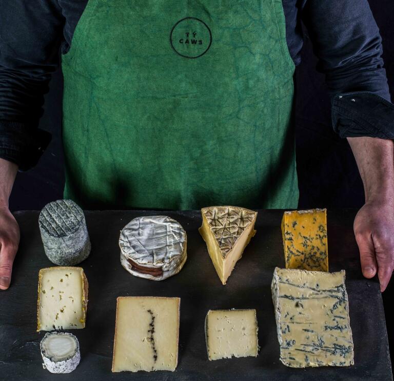 A man holding up a piece of slate with 9 different cheeses on it.