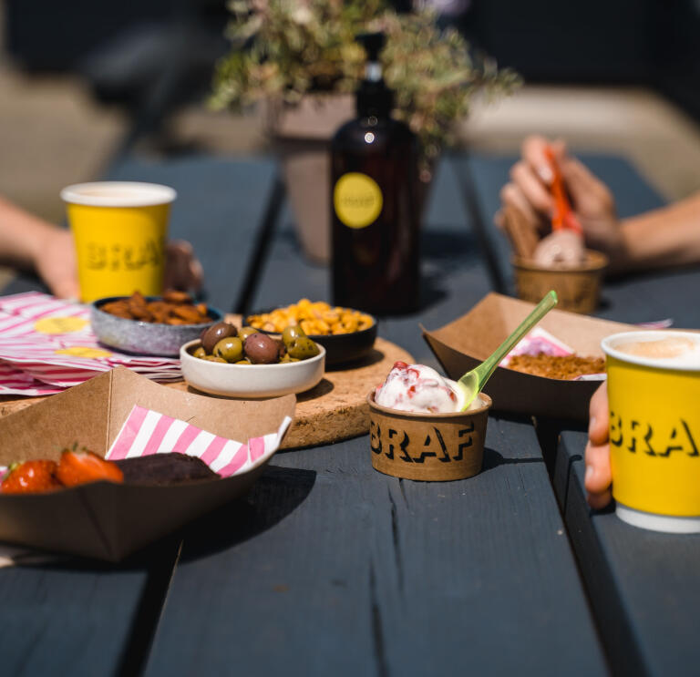 Food and drink on a black wooden bench. There are yellow coffee cups with the word BRAF on them, ice cream, strawberries, olives and nuts.