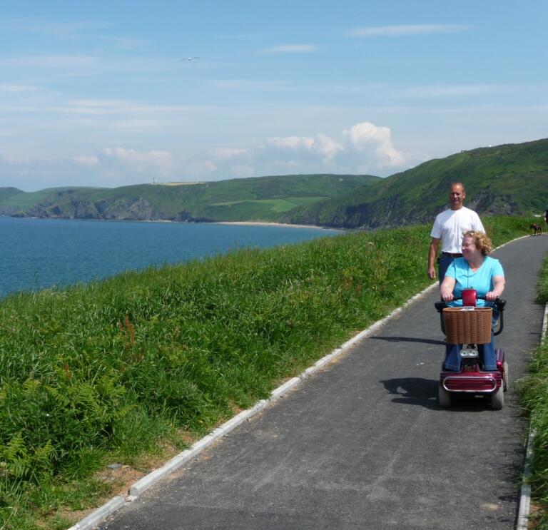 Lady on a mobility scooter with male companion on a tarmac section of the wales coast path looking out to sea