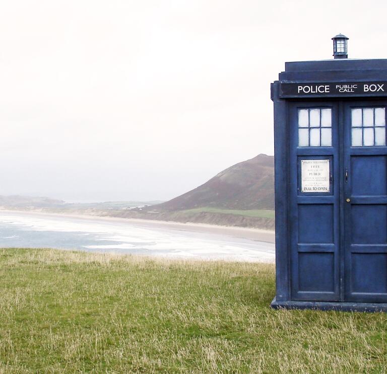 Blue police box film prop on grass with view of vast beach.