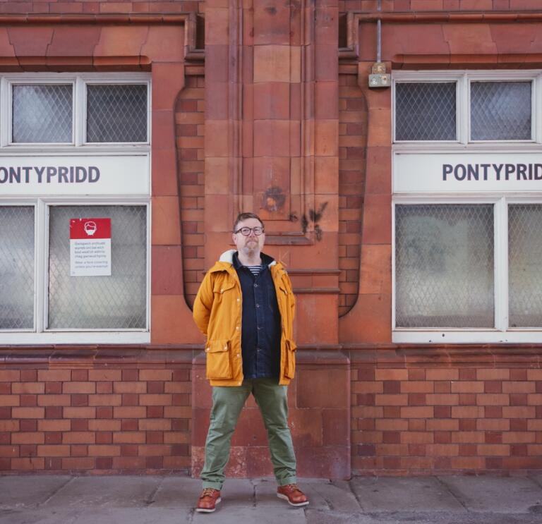 A man in a yellow coat (Gareth Potter_ standing on a train platform. The red brick wall in the background has two white windows with Pontypridd written on them.