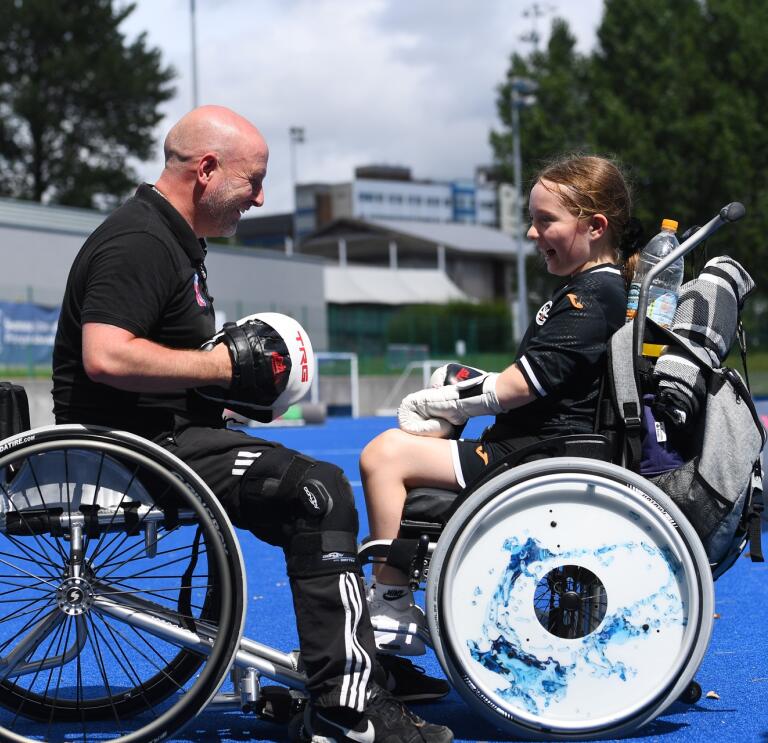 An man and a young girl in wheelchairs facing each other wearing boxing gloves and smiling.
