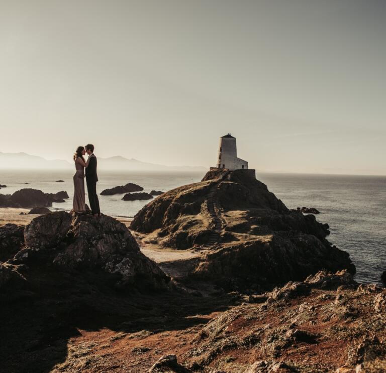 A wedding photograph of a couple kissing on top of a rock on a beach. There is a white lighthouse in the background.