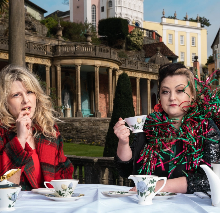 Two women having a cup of tea in ornate gardens.