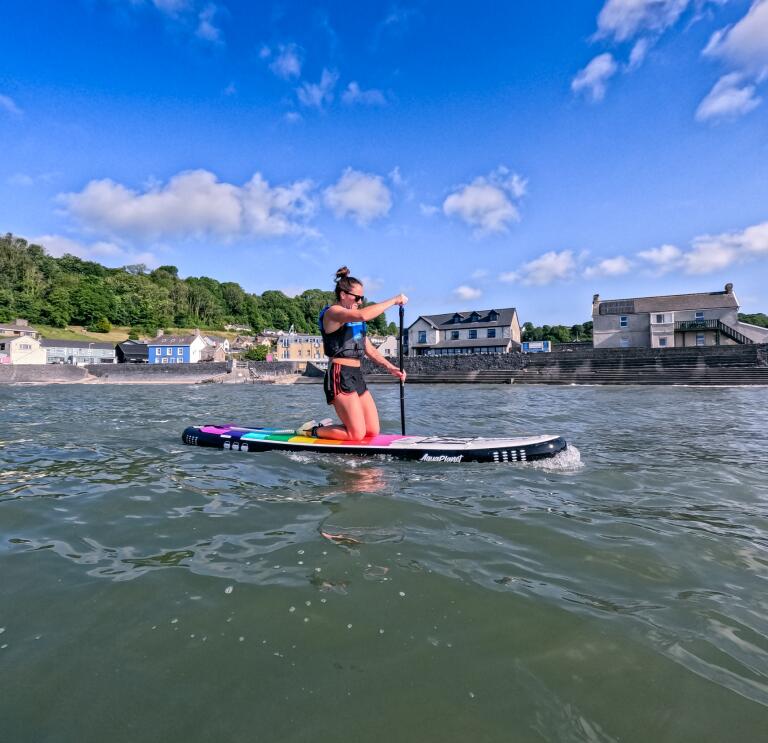 person keelingon a rainbox colour paddleboard on the water, with a row of houses in the background.