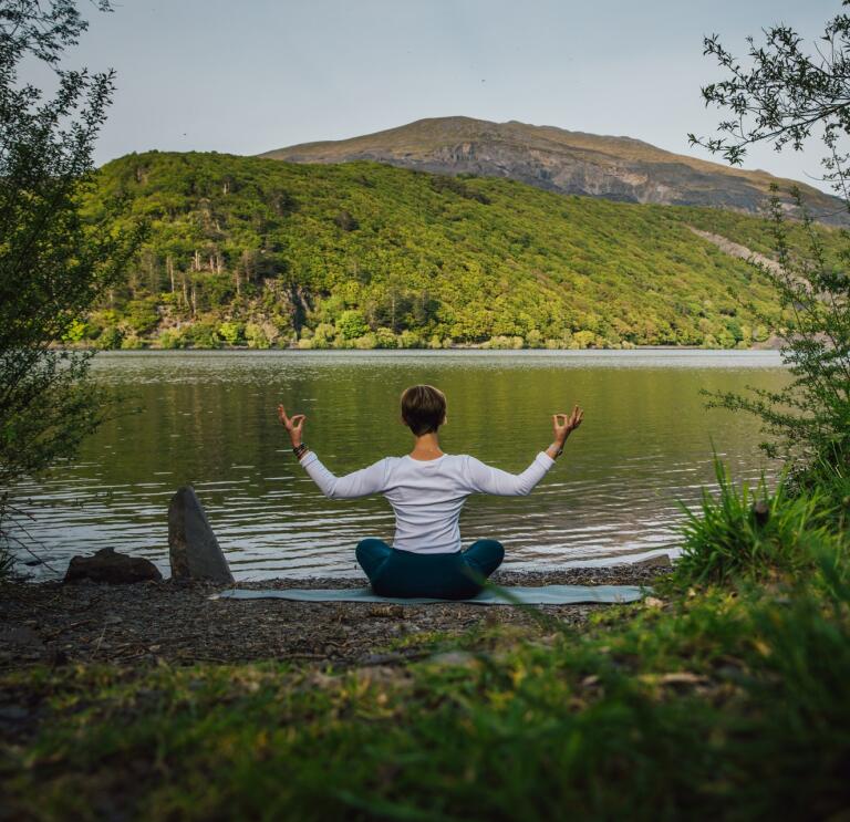 Person seen from behind, sat in a yoga pose in front of a lake with a mountain landscape in the distance