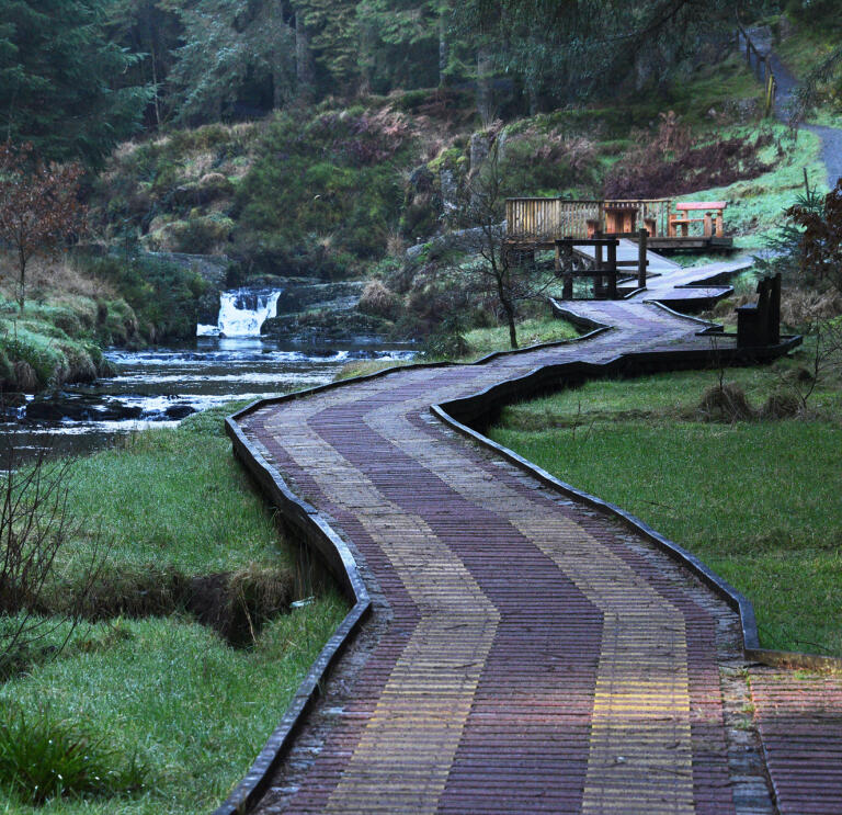 A wide boardwalk in woodland leading to a viewing point, alongside a river.