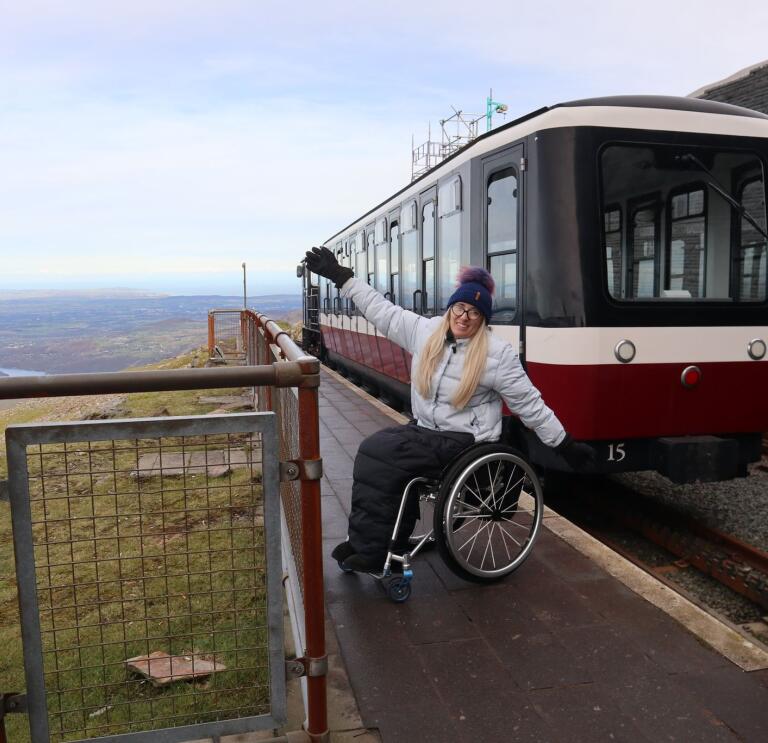 A lady in a wheelchair with arms outstretched by a train at the top of a mountain.