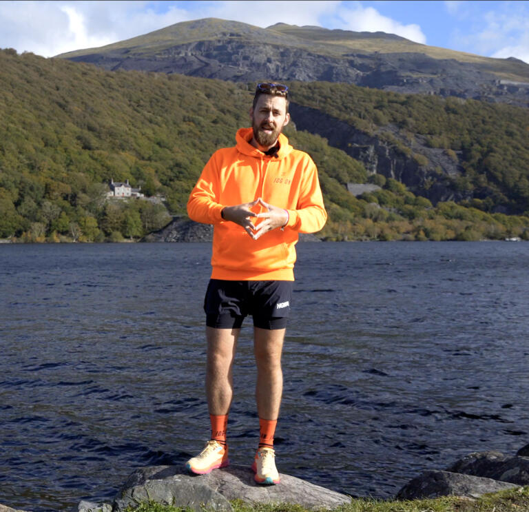 A bearded man in running gear stood on a rock in front of a lake.