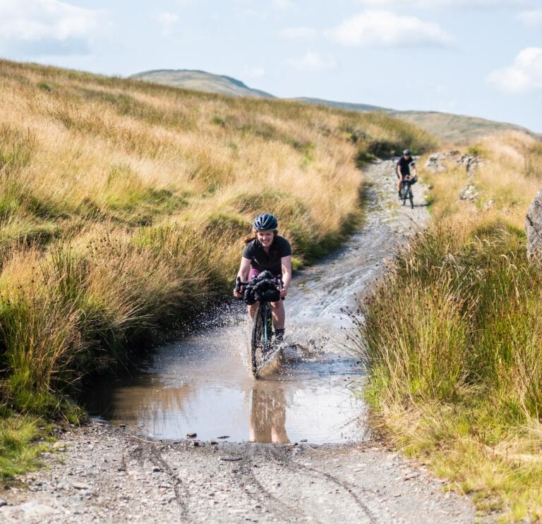 A gravel biker riding through a muddy puddle on a hillside track.