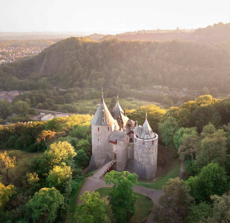 Aerial shot of a fairy tale style castle with pointy turrets and a drawbridge, set amoungst the trees of a woodland. 