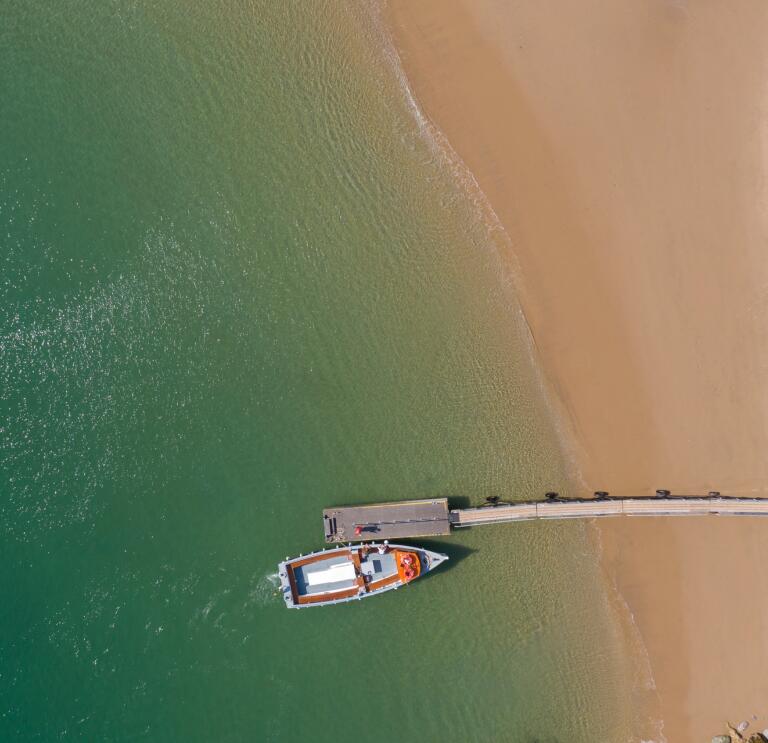 Drone shot of a golden sanded beach from above. There's a red boat moored by a jetty.