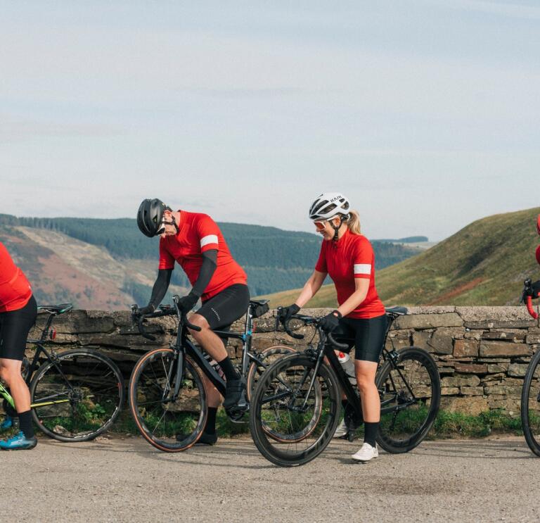 Four cyclists on road bikes by a stone wall overlooking mountains.