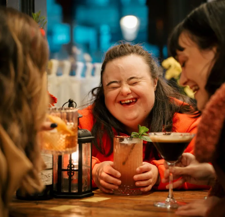 Three people sat around a table with cocktails, the central person is laughing with eyes closed and mouth open.