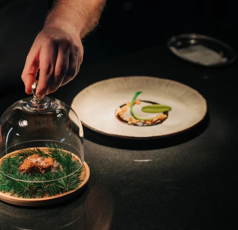 A person lifts a glass dome covering a dish with greens and a small item, next to a plate with arranged food on a dark surface.