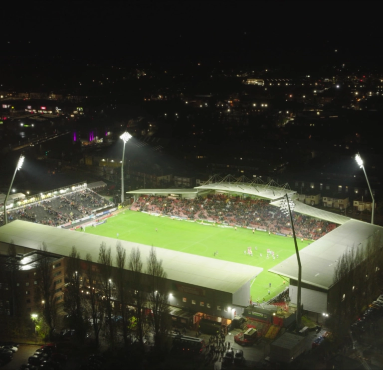 A football stadium in the dark seen from above with flood lights lighting the pitch. THe lights of the surrounding town can be seen in the dark sky around.