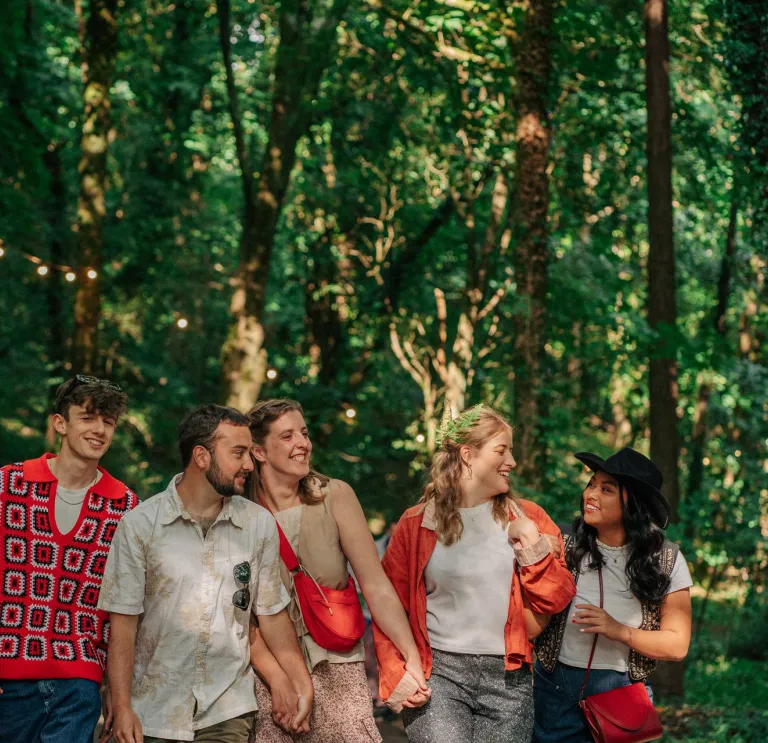 Five individuals strolling in a wooded area under hanging string lights, surrounded by lush greenery and tall trees.
