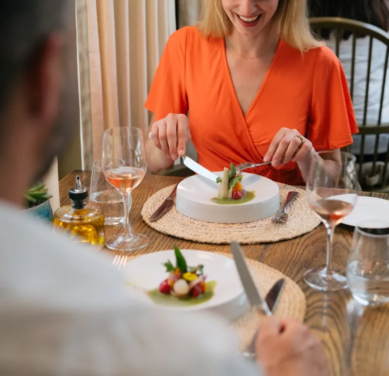 A restaurant scene with two diners, one in focus wearing an orange top, surrounded by food, wine, and tableware.