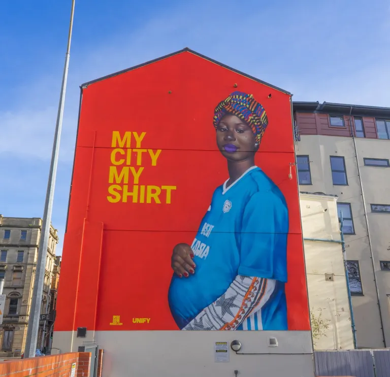 Mural painted on the gable end wall of a building, of a pregnant black woman wearing a colourful headscarf and blue football shirt cradling her baby bump.