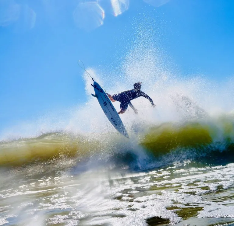 Surfer mid-air above a breaking wave, surrounded by water spray, under a bright blue sky.