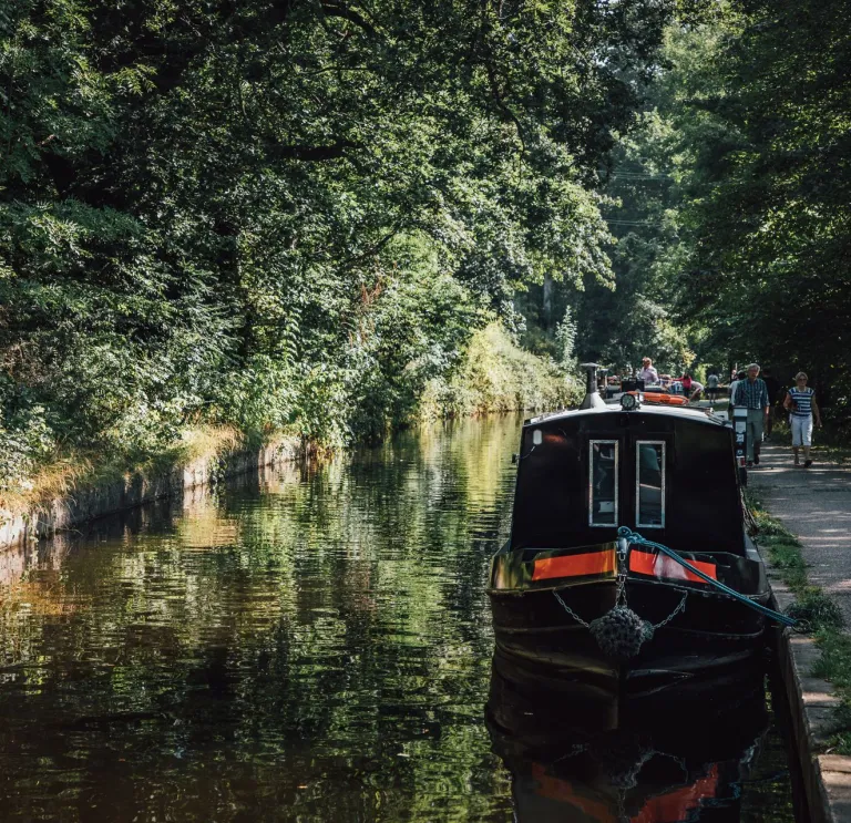 Black and red narrowboat moored on a tranquil canal lined with green trees, with people walking along the adjacent paved path.