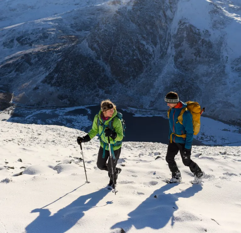 Two hikers walking on a snow-covered mountain summit with backpacks and poles, surrounded by rugged peaks under a partly cloudy sky.