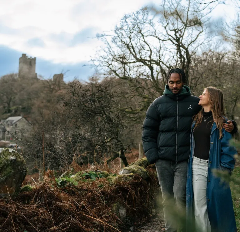 Scenic woodland walk with two people in winter coats, passing a stone castle partially hidden by trees on a hillside.