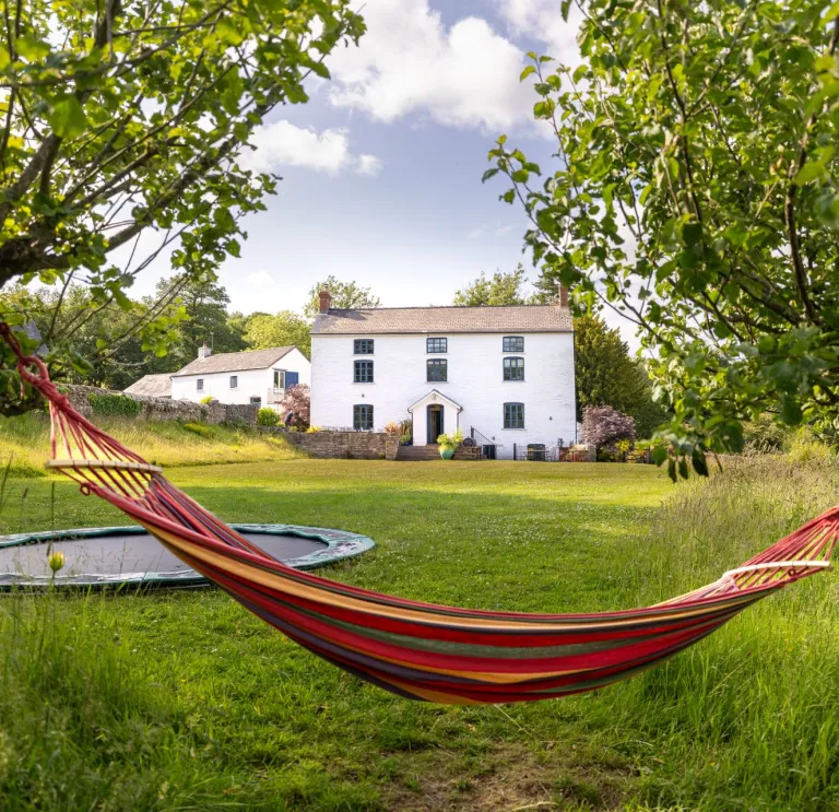Colourful hammock strung between two trees in a grassy garden, with a white farmhouse and trampoline in the background under a blue, partly cloudy sky