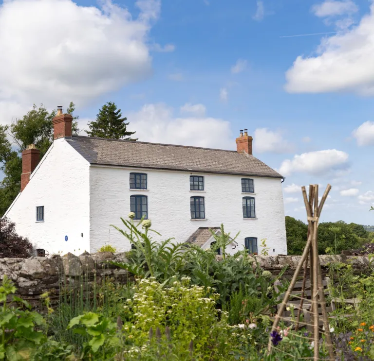 A large, white-washed cottage next to a flower garden. 