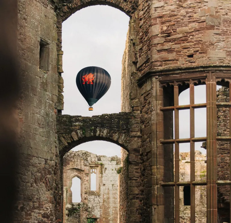 Hot air balloon with a red dragon design floating in the sky, framed by the stone arches and windows of a historic castle ruin.