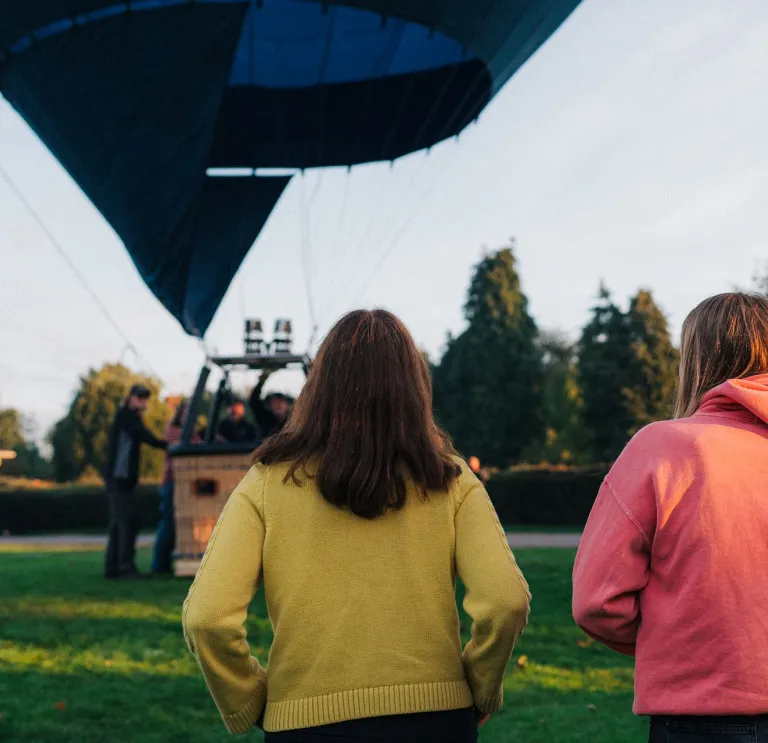 Three people standing on grass watching a hot air balloon being prepared for flight, with trees and a clear sky in the background.