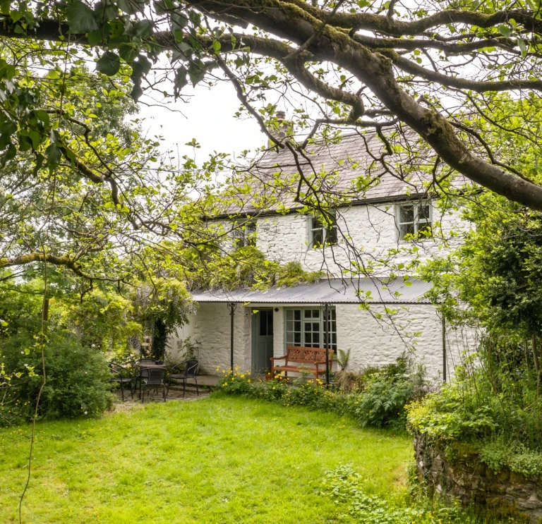 A white painted stone cottage, with a seating area outside, in a lawned garden.