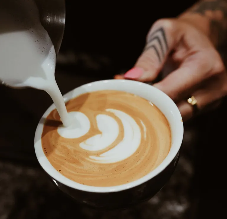 Here’s concise alt text for the second image — and as requested, no mention of any blurred face:  **Alt text:**   *A close-up of steamed milk being poured into a cup of coffee to create latte art and hand with tattoo and ring.