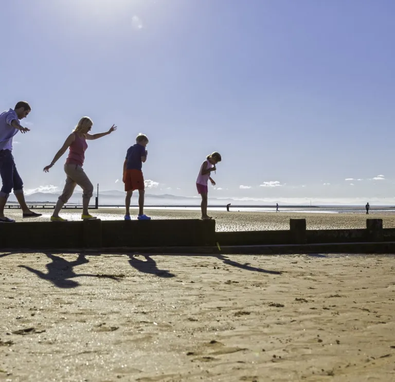 A family walking along a wooden groyne on a sandy beach, with bright sunlight and a clear sky in the background.