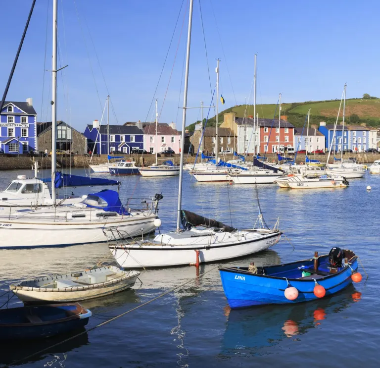 Small boats and yachts moored in a colourful harbour on a sunny day, with brightly painted buildings and green hills in the background.