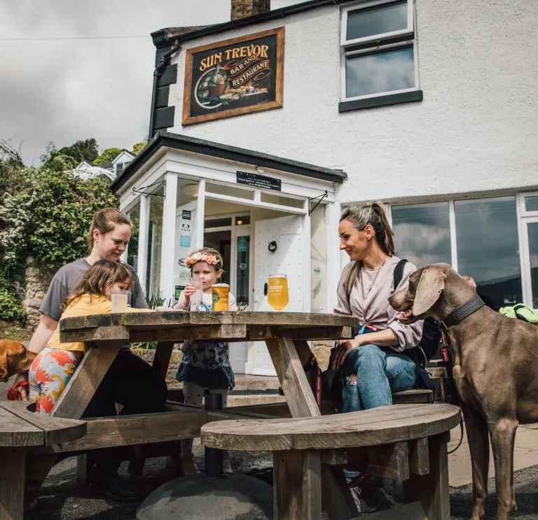 Family sit at a wooden picnic table outside a white pub with drinks, while two dogs stand nearby.