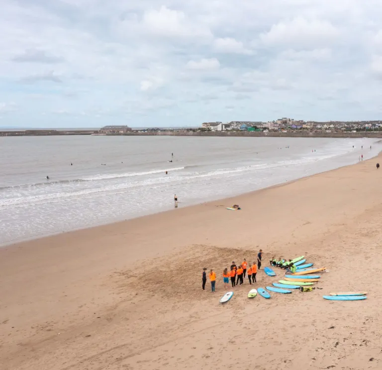 Surf lesson group with surfboards gathered on a wide sandy beach, with gentle waves and a coastal town in the distance.