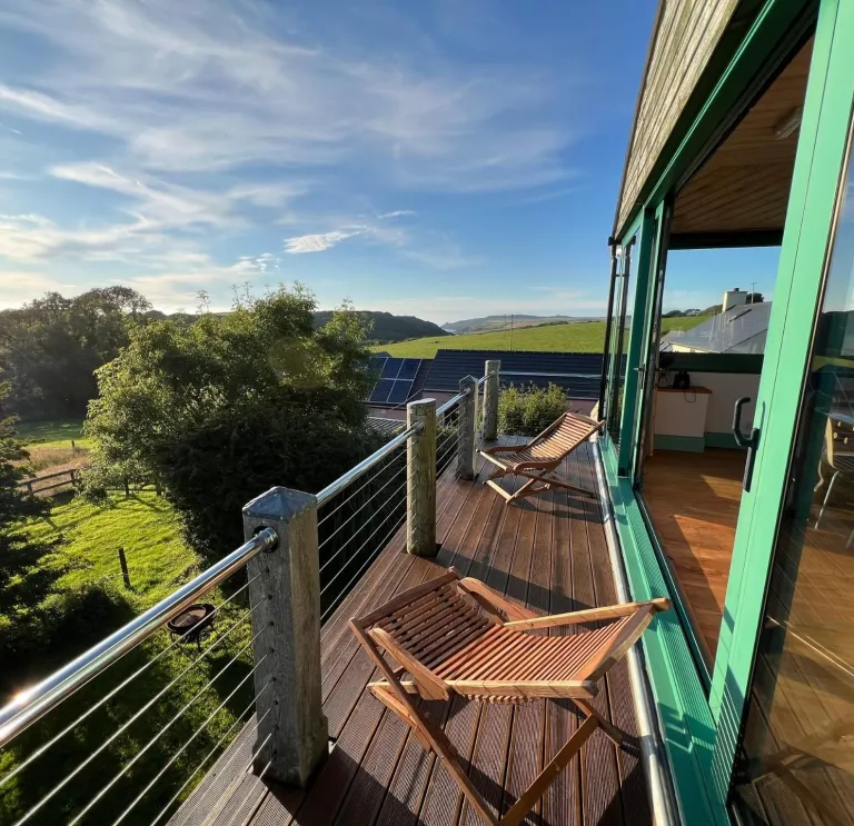 A wooden balcony with folding deckchairs overlooking green countryside, with sunlight streaming across the landscape and large glass doors opening into a modern dining and kitchen area.
